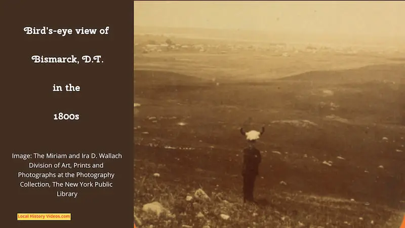 An old photo of a man wearing a buffalo scalp standing on a hillside with Bismarck ND behind him